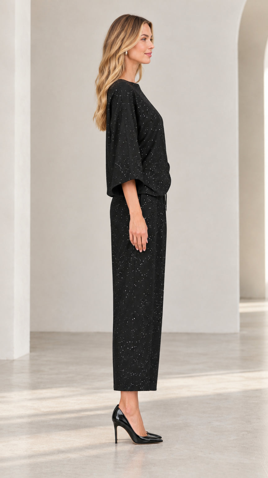 Side profile of model in black shimmer two-piece set featuring 3/4 sleeves and ankle trousers